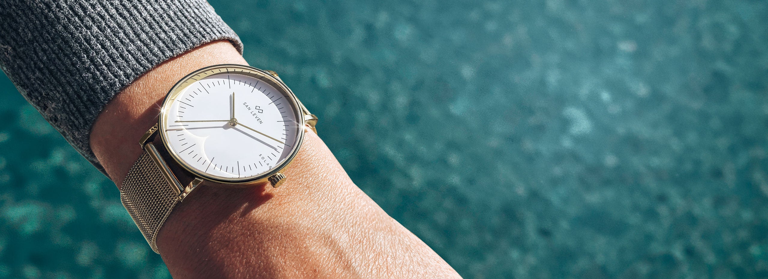 SPARK Solaruhr mit goldenem Mesh-Armband und weißem Zifferblatt am Handgelenk über türkisfarbenem Wasser, Desktop-Ansicht.