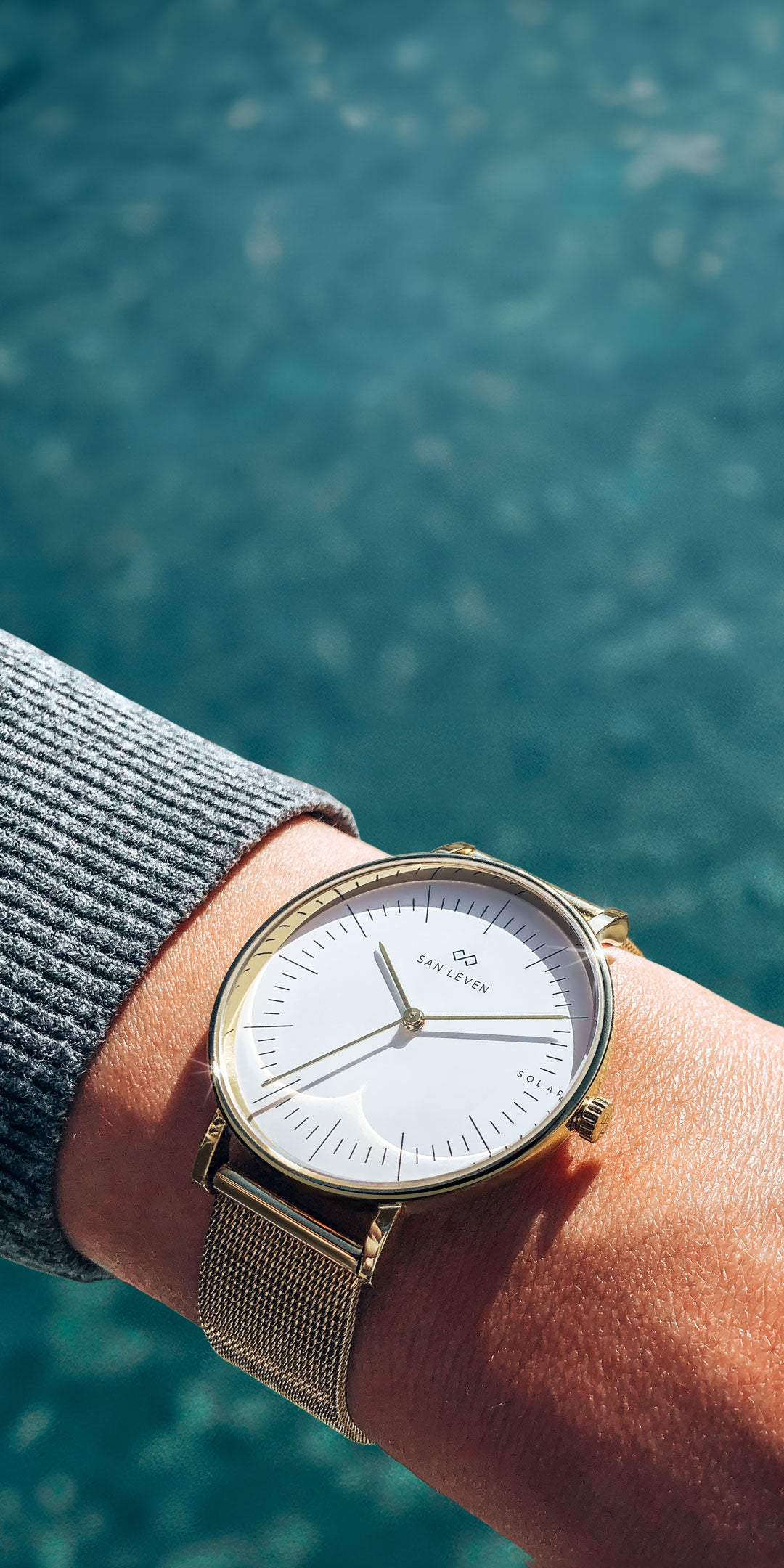 SPARK Solaruhr mit goldenem Mesh-Armband und weißem Zifferblatt am Handgelenk, über Wasser, Mobile-Ansicht.